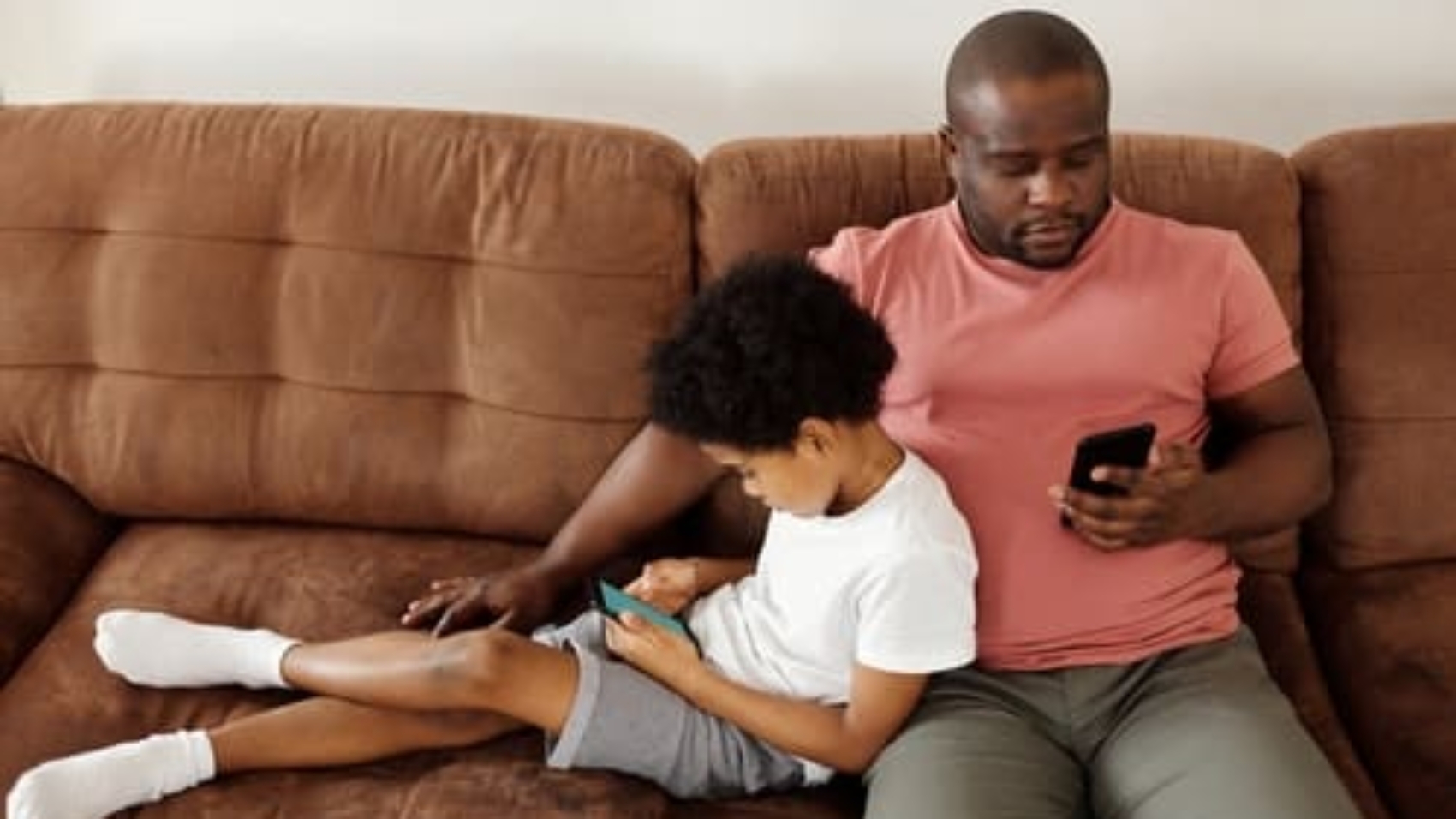 man sitting on couch looking down at cellphone with young child sitting sideways on the couch while leaning against him