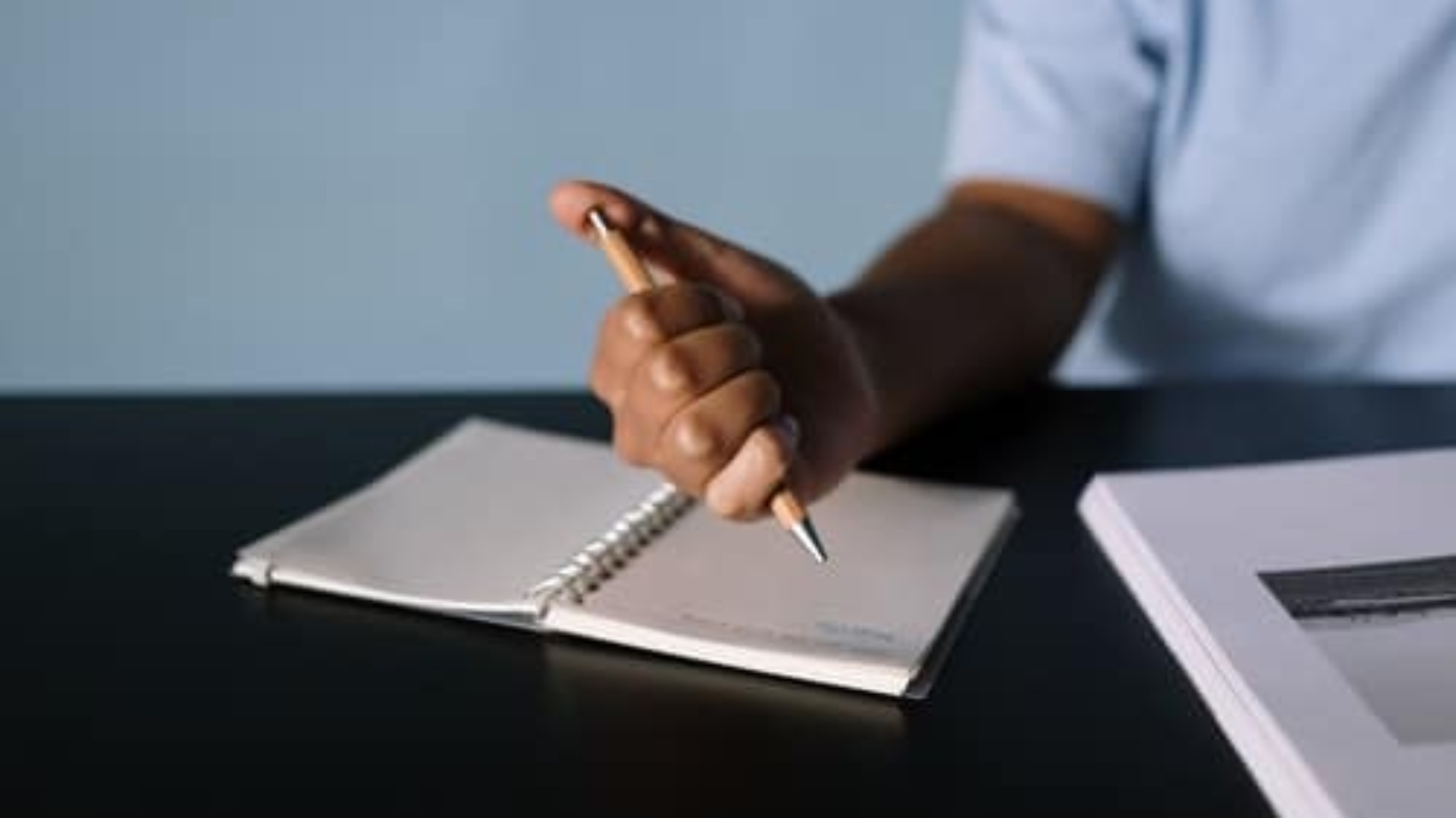 person sitting at desk with hand holding and clicking pen over blank notebook